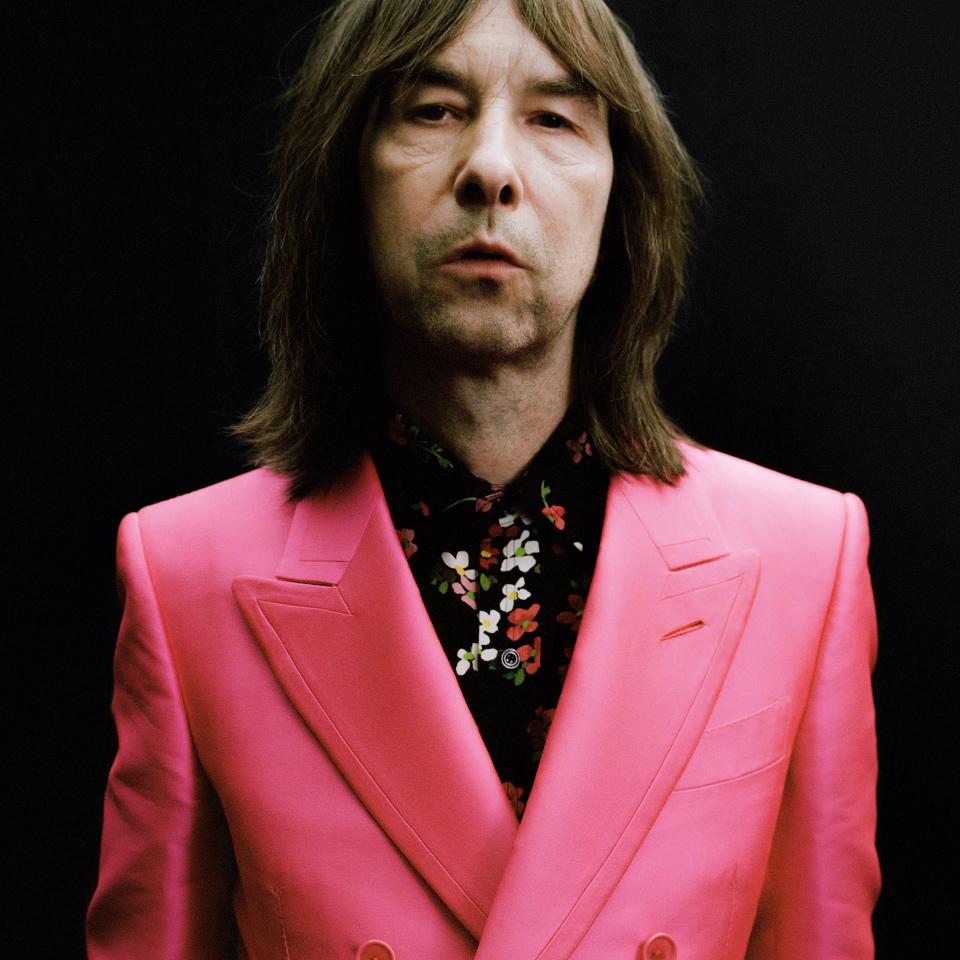 En person med axellångt hår bär en ljusrosa kostymjacka och en svartblommig skjorta och står mot en svart bakgrund.