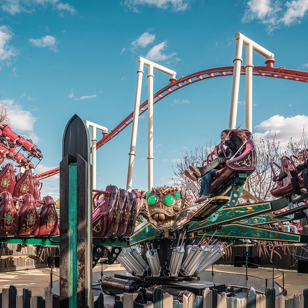 Människor åker på en snurrande nöjesattraktion med säten med insektstema, med en rödvit berg- och dalbana i bakgrunden under en delvis molnig himmel.
