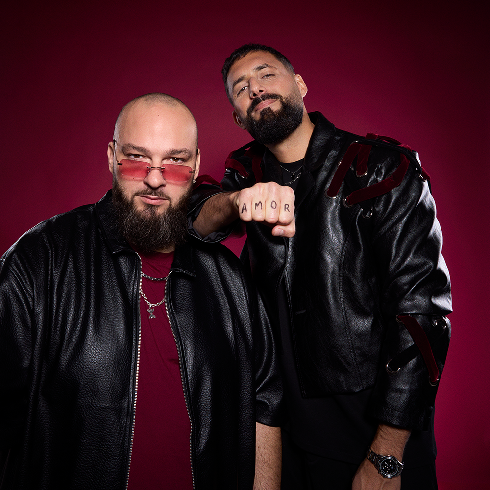 Två män i svarta skinnjackor poserar mot en mörkröd bakgrund; den ene bär rosa glasögon, medan den andre har "AMOR" tatuerat på sina knogar.