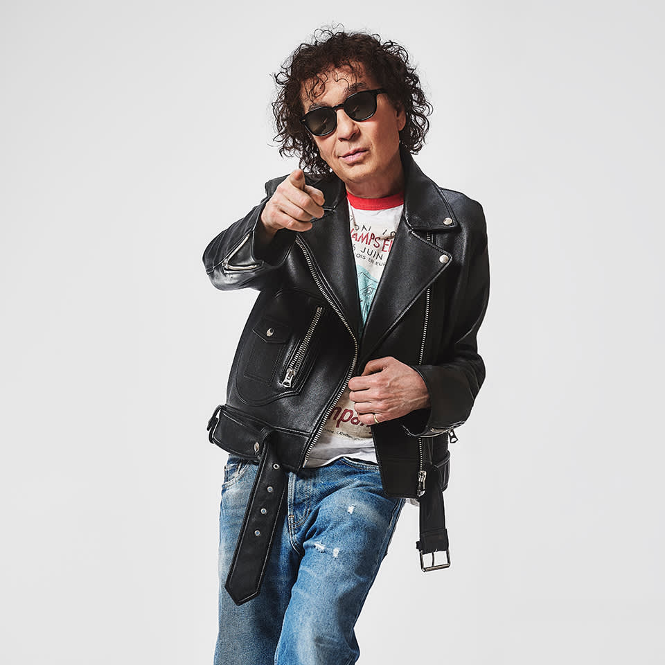 A person with curly hair wearing sunglasses, a leather jacket, and ripped jeans points toward the camera against a plain white background.
