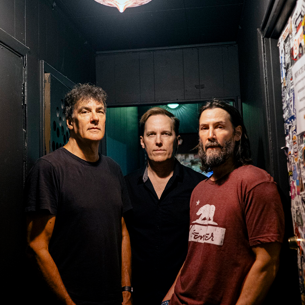 Tre män står sida vid sida i en svagt upplyst korridor, vända mot kameran. En av dem bär en svart skjorta, en annan en mörk knappskjorta och den tredje en röd t-shirt med björnmotiv.