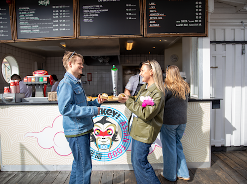 Two people smiling and holding food trays stand in front of a food counter with a monkey graphic, while another person faces the menu above.