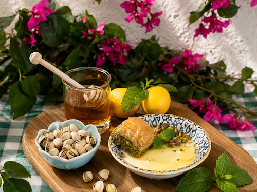 A plate with baklava, pistachios, and syrup, a bowl of pistachios, a glass of honey, lemons, and mint on a tray, set on a checkered cloth with flowers in the background.