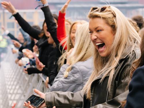 En grupp människor står bakom en metallbarriär vid ett utomhusevenemang, med en kvinna framför som skrattar och håller i en telefon.