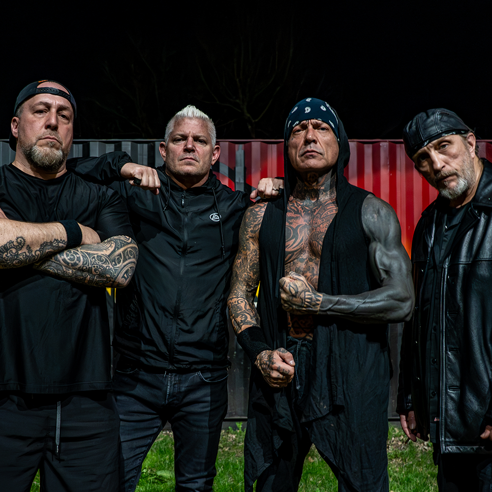 Fyra män med tatueringar och allvarliga ansiktsuttryck poserar utomhus på kvällen framför en röd och svart fraktcontainer.