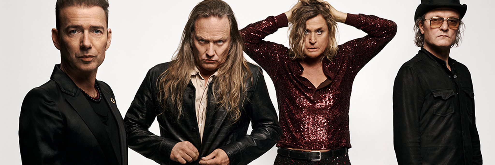 Fyra män står mot en vit bakgrund, iklädda mörka och mönstrade kläder, med varierande ansiktsuttryck och poser.