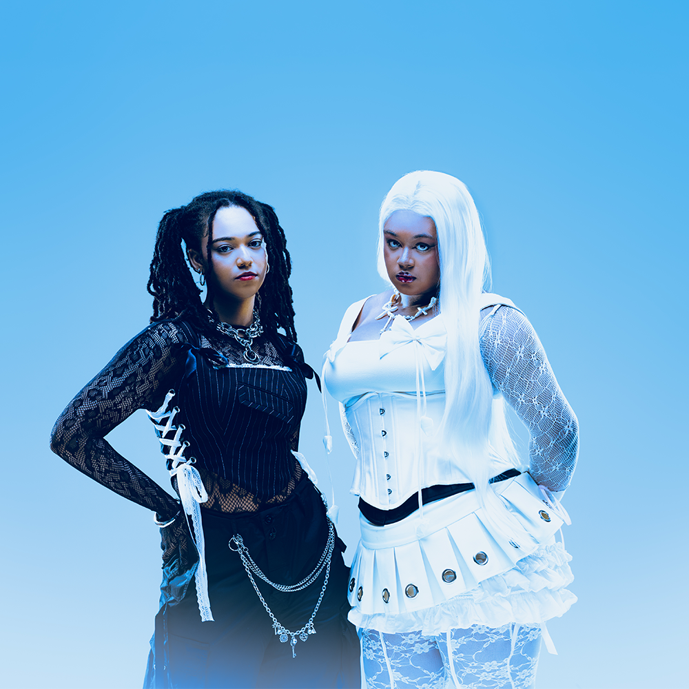 Två kvinnor står sida vid sida mot en blå bakgrund, den ena svartklädd med dreadlocks, den andra vitklädd med långt rakt hår och veckad kjol.