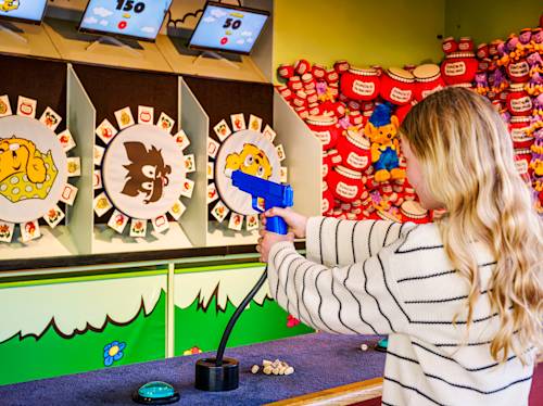 A person with long blonde hair aims a toy gun at targets in a carnival shooting game, with plush prizes displayed in the background.
