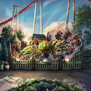 People sit in a circular amusement park ride themed as a giant mechanical spider, surrounded by a white fence, with a roller coaster track visible in the background.
