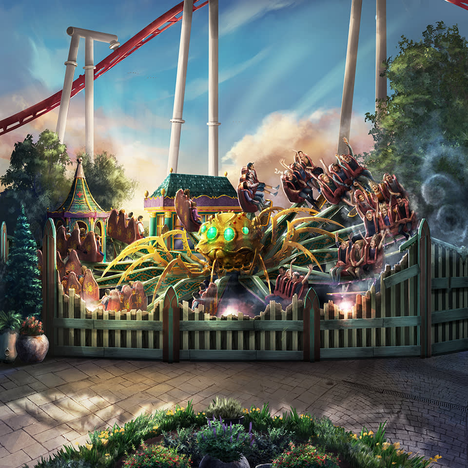 People sit in a circular amusement park ride themed as a giant mechanical spider, surrounded by a white fence, with a roller coaster track visible in the background.