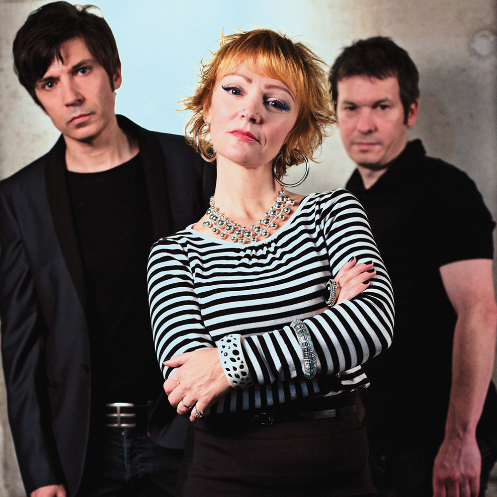 Tre personer poserar för ett foto; en kvinna i randig skjorta står med armarna i kors i förgrunden, två män står bakom henne, alla vända mot kameran.