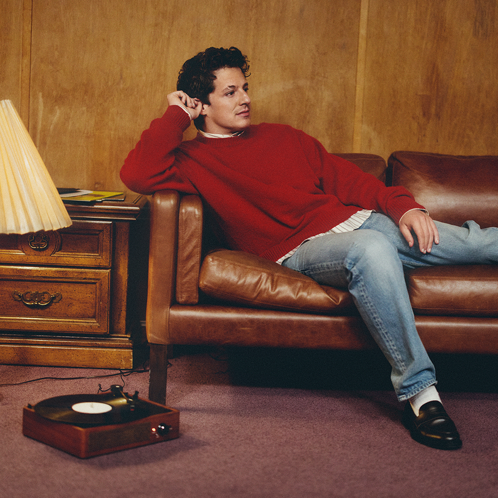 En man i röd tröja och jeans ligger i en brun skinnsoffa bredvid ett sidobord i trä med en lampa; en skivspelare står på mattan intill.