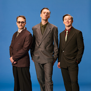 Tre kostymklädda män står sida vid sida mot en blå bakgrund och vänder sig mot kameran med neutrala eller lätt leende ansiktsuttryck.