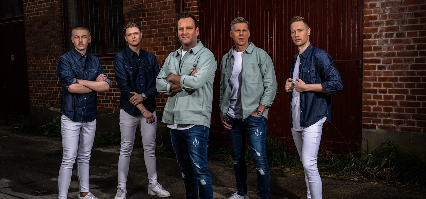 Fem män poserar utomhus mot tegelväggar, iklädda denim och ljusa jackor med vita byxor, i en stadsmiljö.