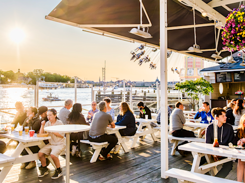 Människor sitter vid vita picknickbord på en terrass vid floden och äter och pratar i solen. I bakgrunden syns nöjesfält, vatten och en stadsbild.