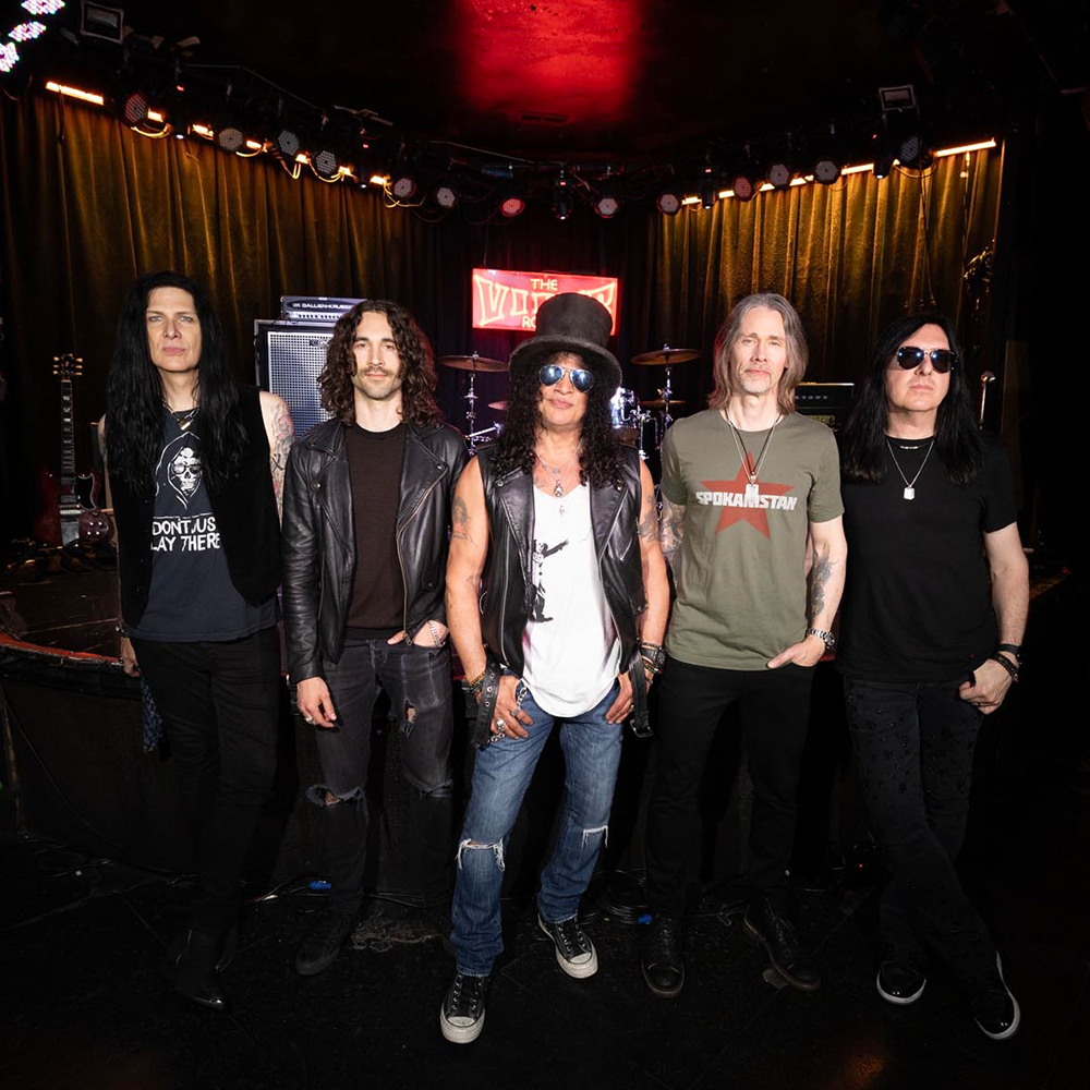 Five men stand together on a stage with musical equipment and a red neon sign reading “The Viper Room” in the background.