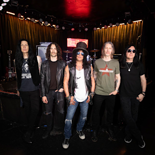 Five men stand together on a stage with musical equipment and a red neon sign reading “The Viper Room” in the background.