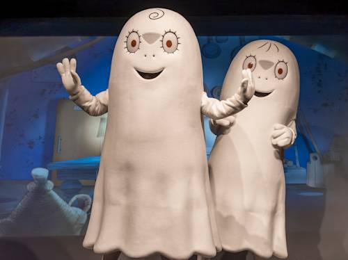 Two people in large white ghost-like costumes with cartoon faces stand indoors, waving, against a blue-themed background.