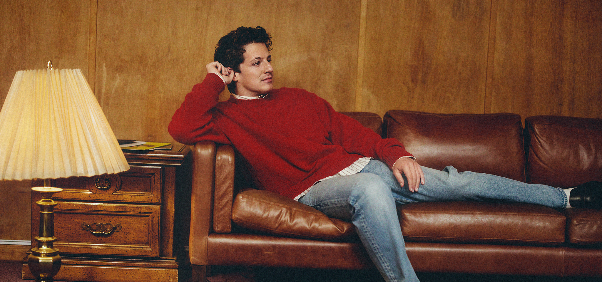 En person i röd tröja och jeans ligger i en brun skinnsoffa bredvid ett sidobord i trä med en lampa.
