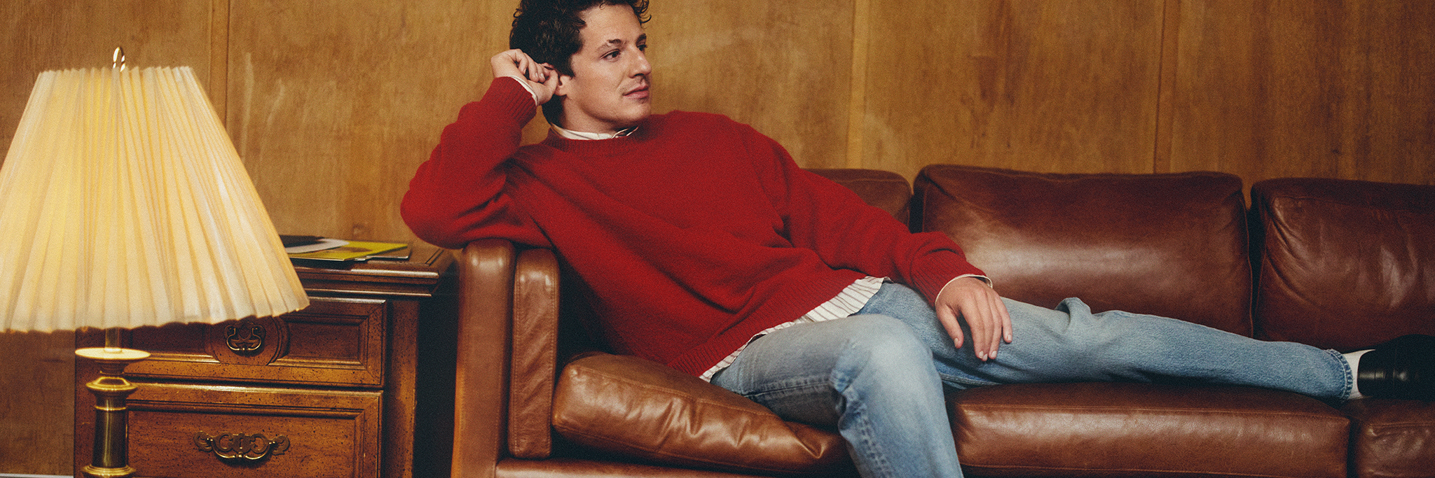 En person i röd tröja och jeans ligger i en brun skinnsoffa bredvid ett sidobord i trä med en lampa.