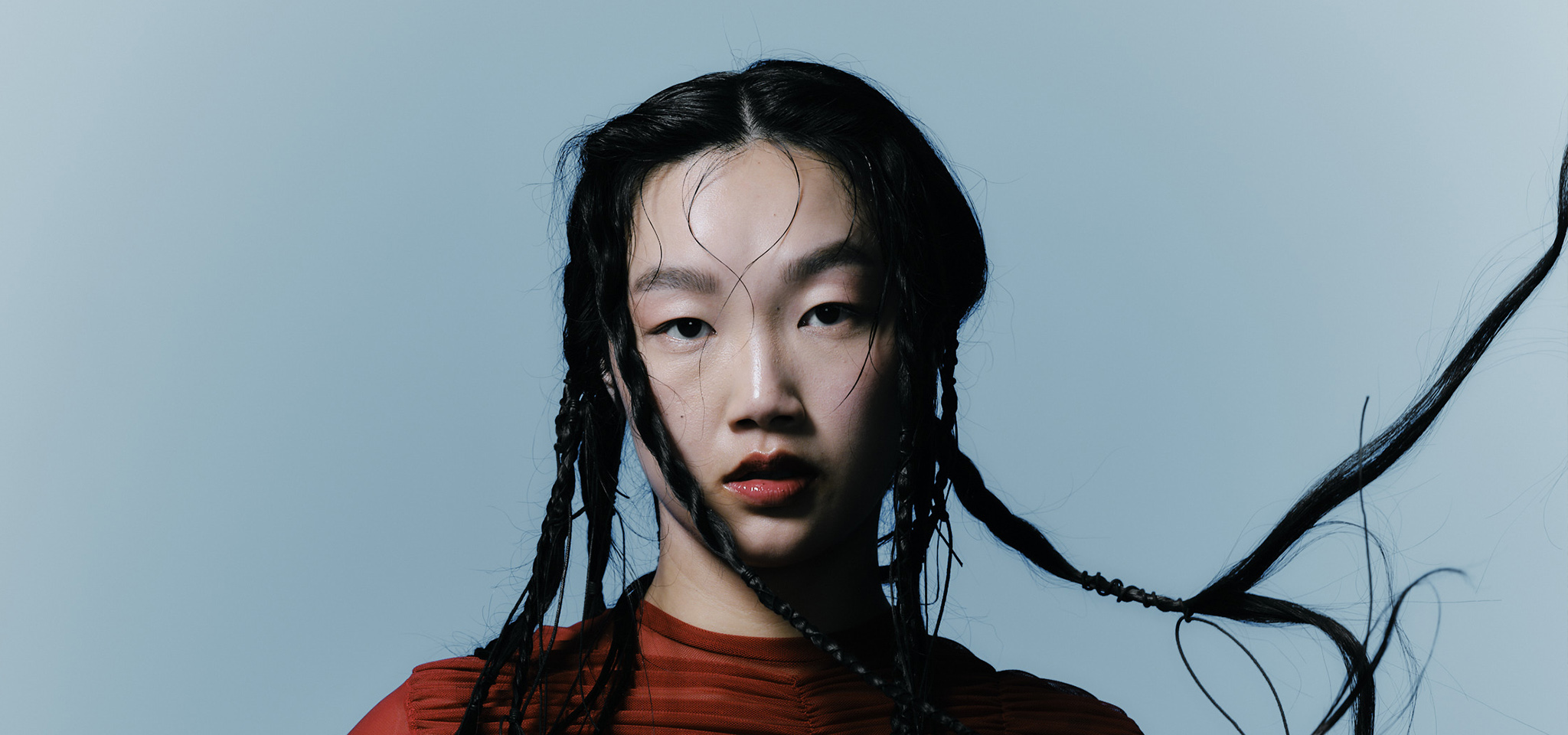 En person med långt flätat hår och röd topp tittar direkt in i kameran mot en ljusblå bakgrund.