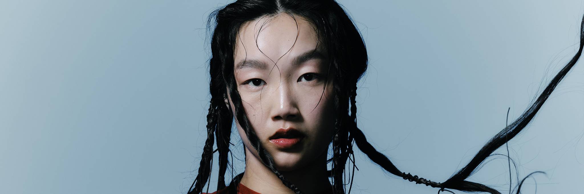 En person med långt flätat hår och röd topp tittar direkt in i kameran mot en ljusblå bakgrund.