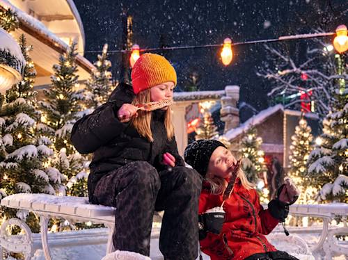 Två barn i vinterjackor som sitter på en snötäckt bänk och äter chokladöverdragna våfflor på pinnar i en julig utomhusmiljö.
