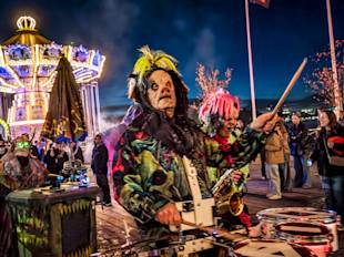 Människor i färgglada, kusliga dräkter och masker spelar musikinstrument i en festlig utomhusmiljö med en karusell och åskådare i bakgrunden.