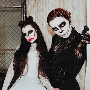Två personer i detaljerade svartvita halloweensminkningar och dräkter poserar inomhus, den ena i en vit klänning med mörk ögonmakeup, den andra i en svart dräkt med en dödskalle i ansiktet.