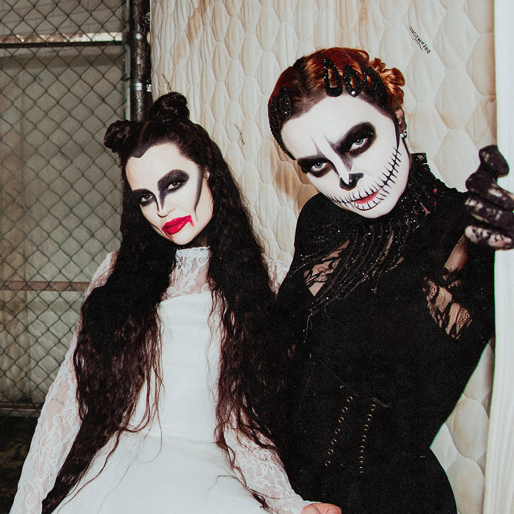 Två personer i detaljerade svartvita halloweensminkningar och dräkter poserar inomhus, den ena i en vit klänning med mörk ögonmakeup, den andra i en svart dräkt med en dödskalle i ansiktet.