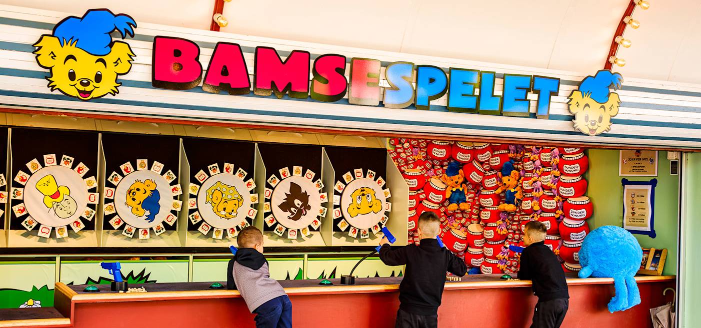 Three children play a carnival-style game called "Bamsespelet," featuring colorful character wheels and plush toy prizes.