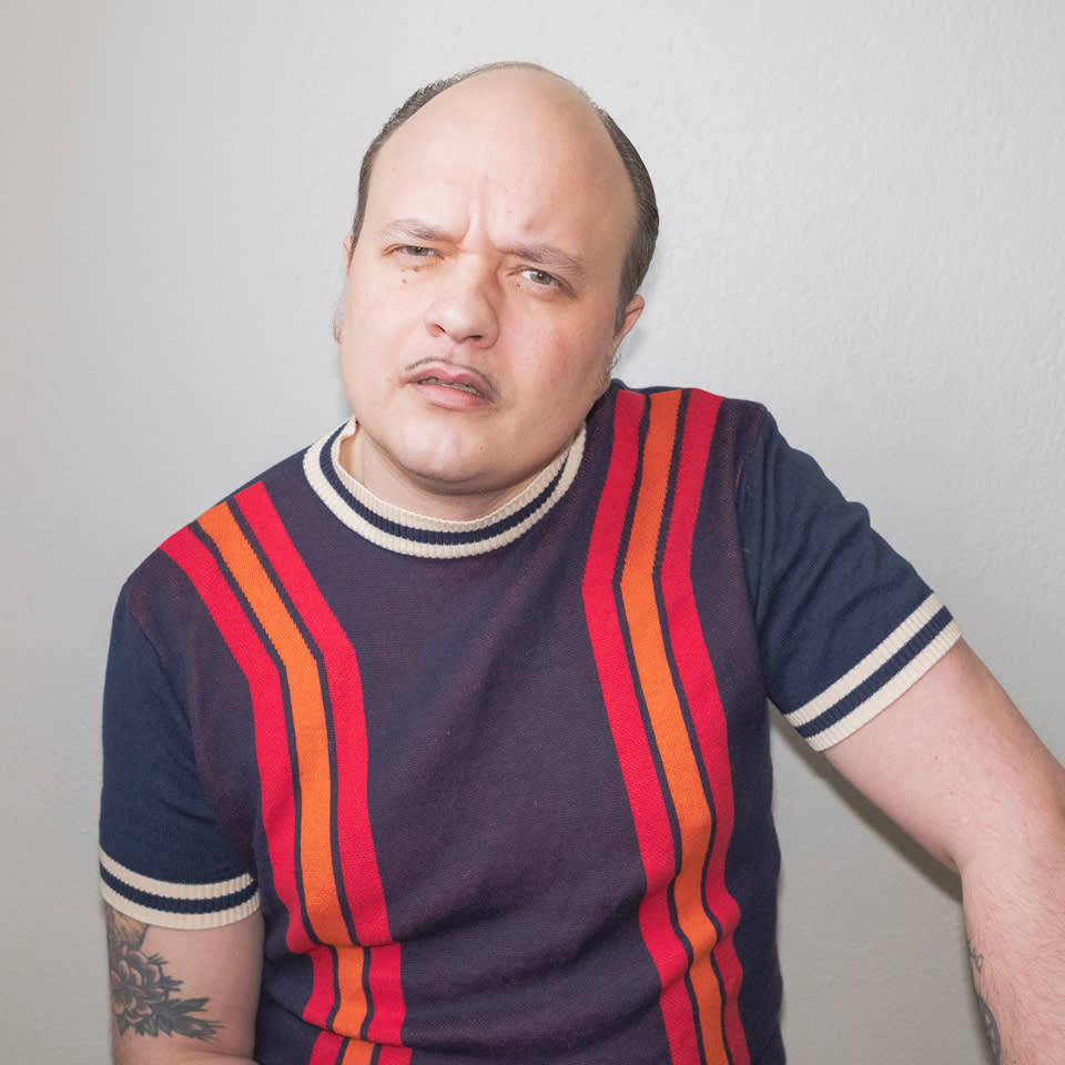 En man med tillbakadraget hårfäste och tatuerad arm bär en marinblå skjorta med röda och orange ränder och poserar mot en enfärgad ljus vägg.