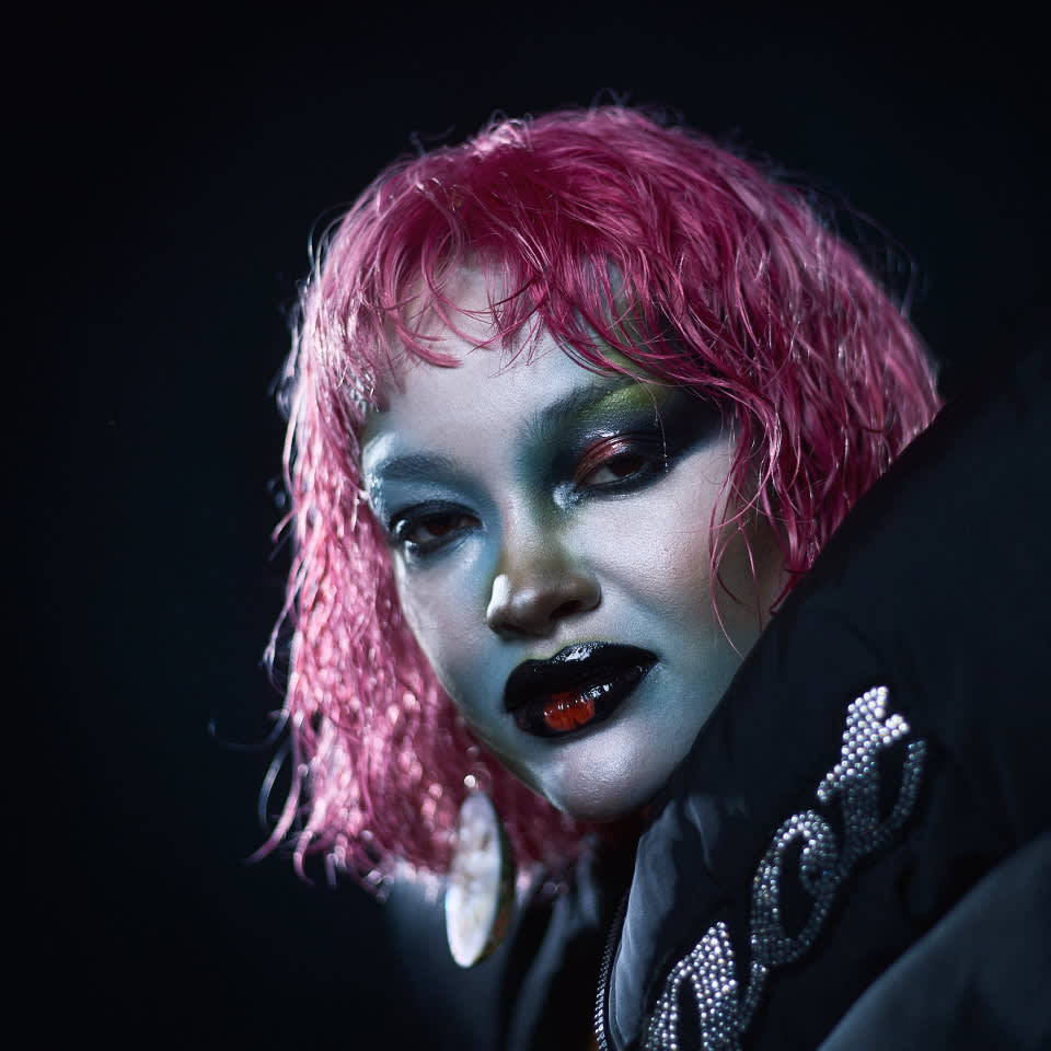 Person med kort rosa hår, dramatisk metallic makeup och glansiga svarta läppar, klädd i en mörk jacka med glittrande utsmyckningar, mot en mörk bakgrund.