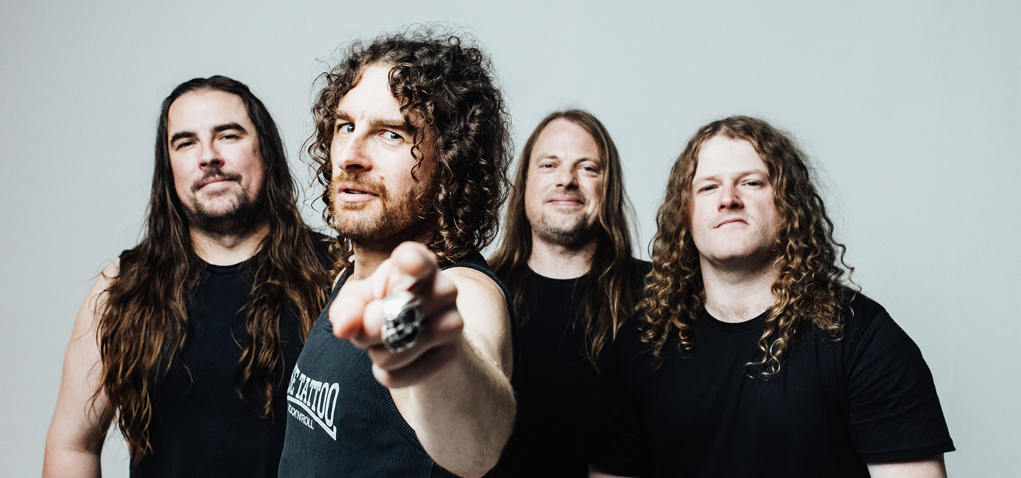 Fyra män med långt hår och svarta skjortor står tillsammans; en man längst fram pekar mot kameran medan de andra vänder sig framåt och ler.