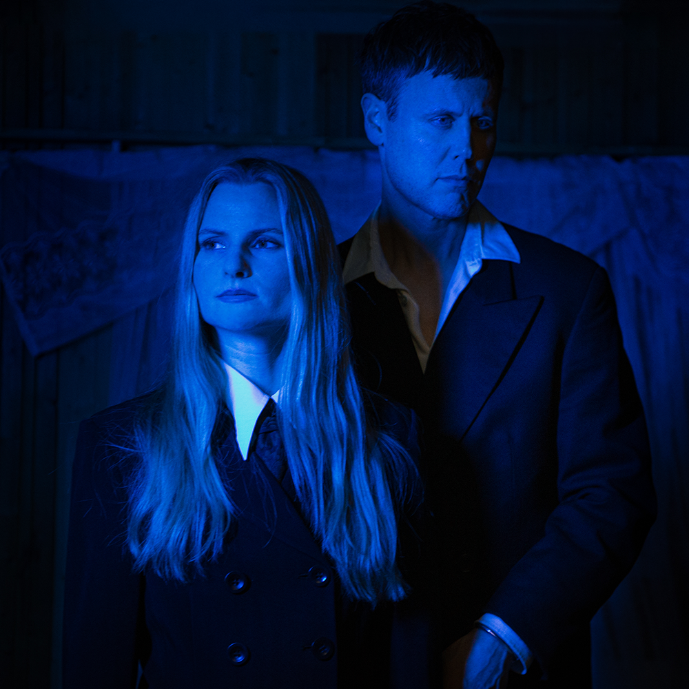Two people in dark formal clothing stand side by side under dramatic blue lighting, looking in different directions against a dim indoor background.