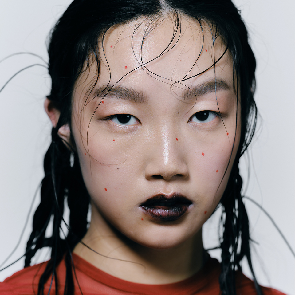 Närbild av en person med blött hår, mörkt läppstift och flera små röda prickar i ansiktet, klädd i röd topp, mot en enfärgad bakgrund.