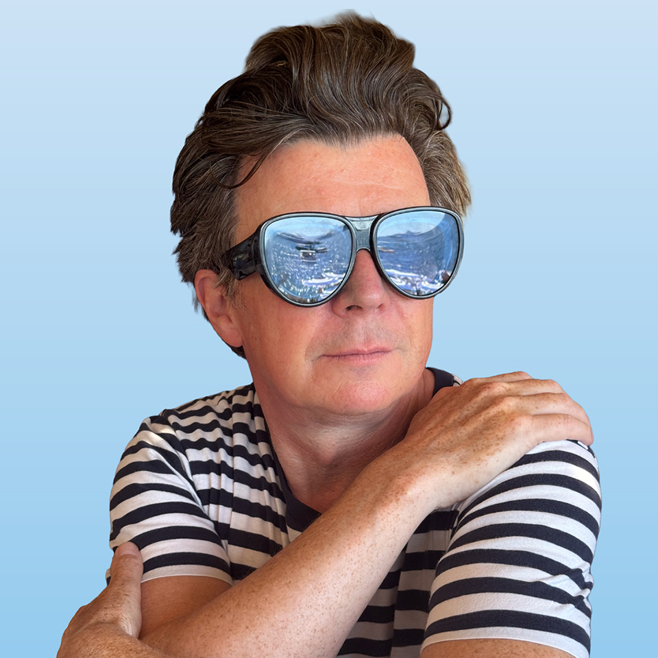 En person med stora reflekterande solglasögon och en svartvitrandig skjorta sitter med ena armen vilande på axeln mot en ljusblå bakgrund.