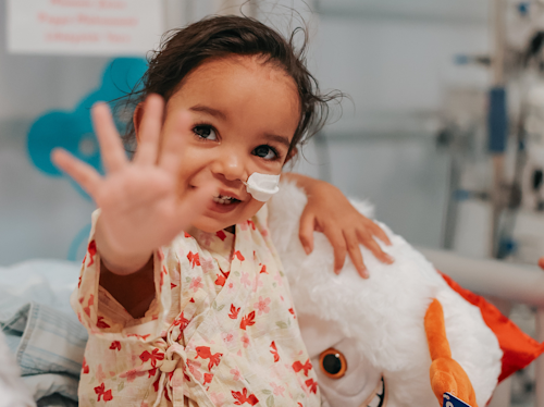 Ett litet barn i sjukhuskläder med nässlang ler och räcker upp handen, sittande bredvid en gosedjur i en medicinsk miljö.