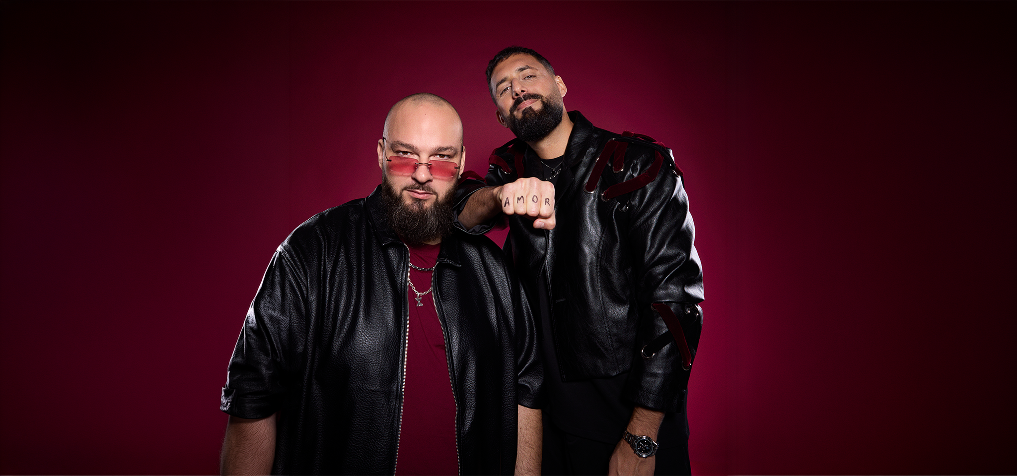 Två män poserar mot en djupröd bakgrund, båda klädda i svarta skinnjackor; den ene bär tonade glasögon och den andre har "AMOR" tatuerat på sina fingrar.