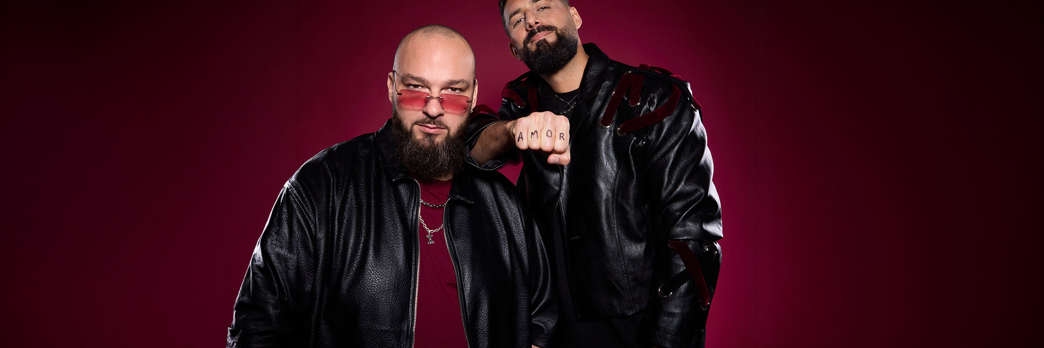 Två män poserar mot en djupröd bakgrund, båda klädda i svarta skinnjackor; den ene bär tonade glasögon och den andre har "AMOR" tatuerat på sina fingrar.