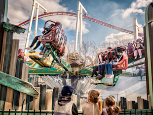 Three children watch as people ride a spinning amusement park attraction with decorative seats, under a partly cloudy sky with a roller coaster in the background.