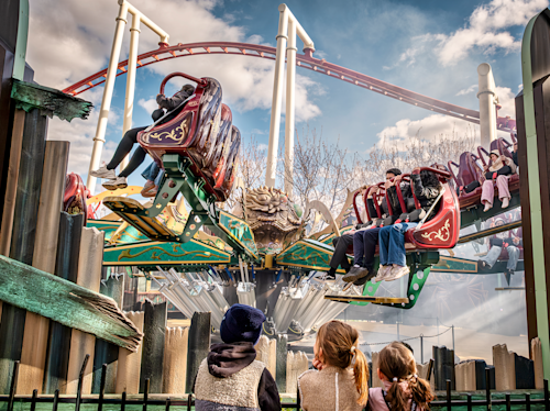 Tre barn tittar på en snurrande åkattraktion på ett nöjesfält med människor i röda och gröna bilar, med berg- och dalbanor i bakgrunden under en molnig himmel.