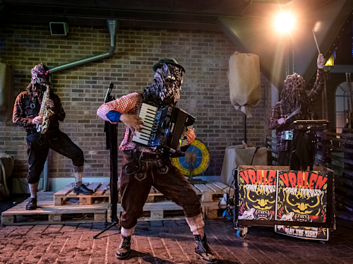 Tre musiker i monstermasker spelar saxofon, dragspel och keyboard på en liten scen med tegelvägg som bakgrund och rekvisita, bredvid en resväska märkt "Vralen Spook Straat".