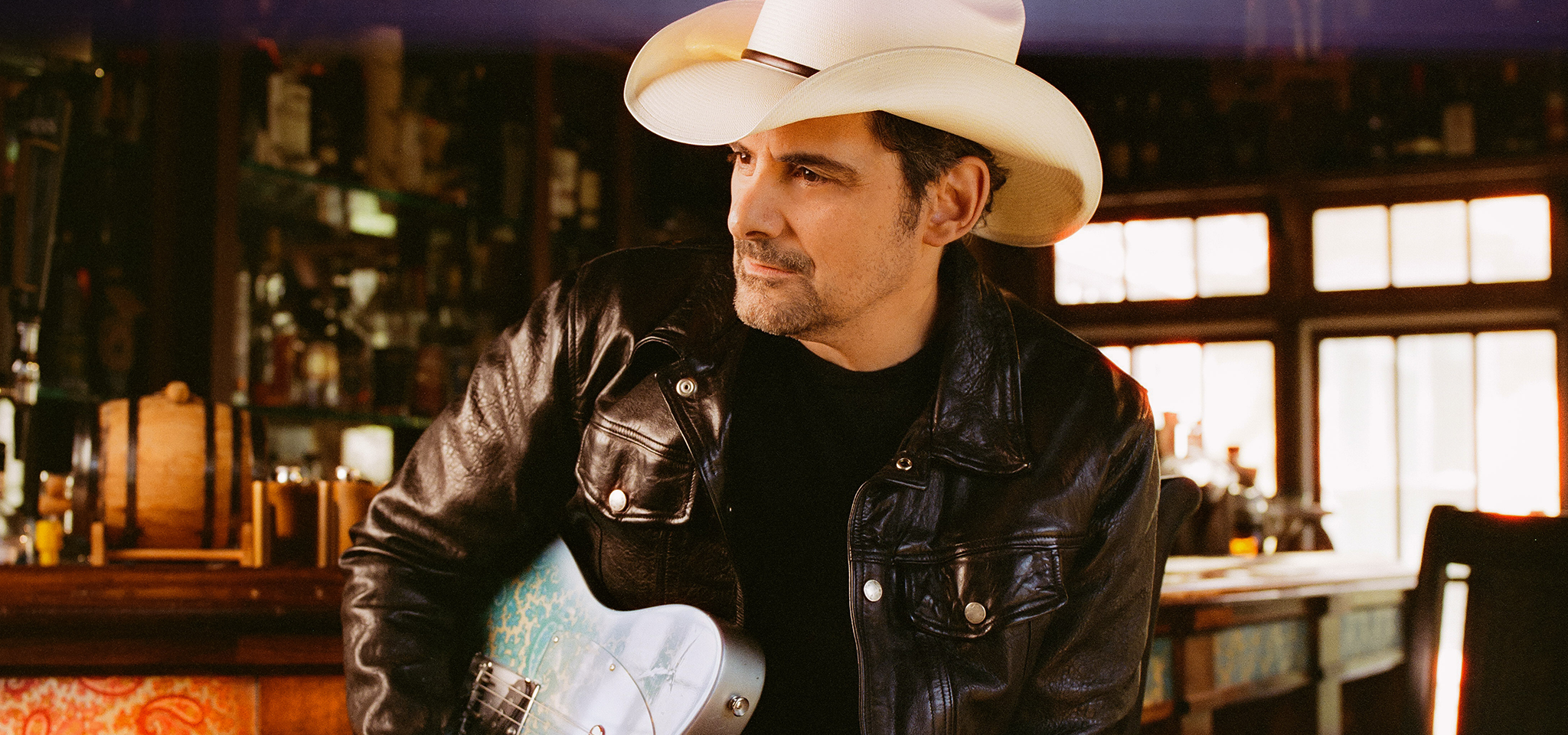 En man i svart skinnjacka och vit cowboyhatt håller i en elgitarr medan han sitter i en bar.