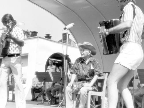 Three musicians perform on an outdoor stage, one playing guitar, one playing accordion, and one seated in the center wearing a cowboy hat; other musicians sit in the background.