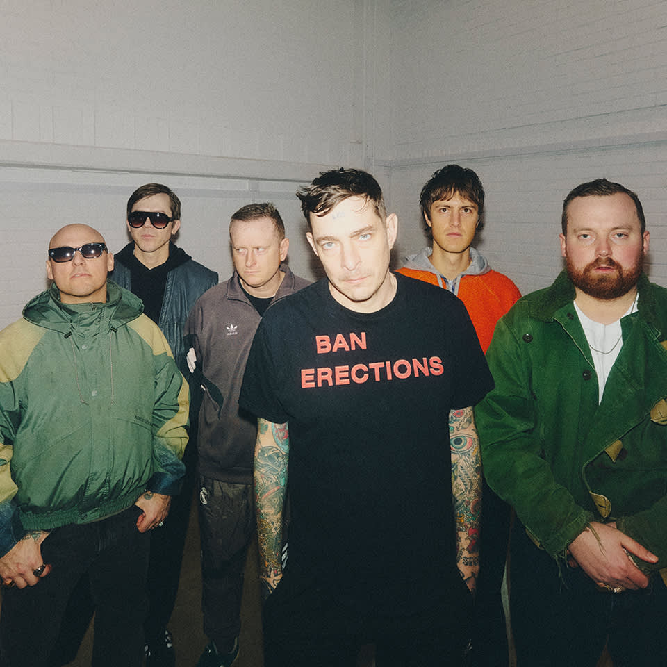Six men stand indoors against a white brick wall; the central figure wears a black t-shirt with the text "BAN ERECTIONS.