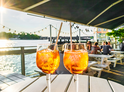Två Aperol Spritz-cocktails på ett bord i en utomhusbar vid floden, med solljus, vatten, ljusslingor och människor i bakgrunden.