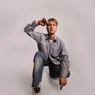 Person som ligger på knä, iklädd en rutig skjorta och jeans, tittar på kameran mot en vanlig bakgrund.
