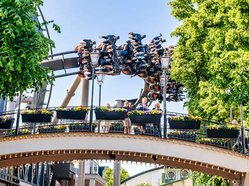 People ride an inverted roller coaster above a bridge with onlookers and flower boxes, surrounded by green trees on a sunny day.