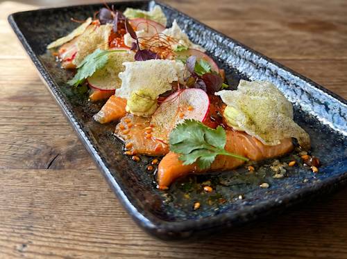 A rectangular black plate with thinly sliced sashimi, topped with radish, herbs, crisp garnishes, and a drizzle of sauce on a brown textured surface.